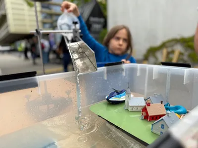 Kind gießt Wasser mit einer Schaufel in einen durchsichtigen Behälter mit einem Modell von Häusern und einer Kirche auf grünem Untergrund