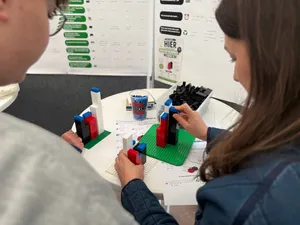 Zwei Personen bauen mit bunten Bausteinen auf grünen Bauplatten an einem weißen Tisch, im Hintergrund Informationsplakate