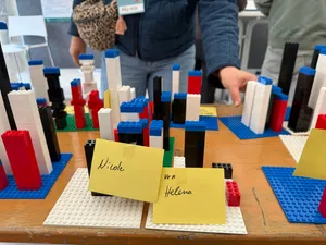 Mehrere bunte Lego-Türme auf weißen und blauen Grundplatten mit gelben Zetteln, auf denen 'Nicole' und 'Helena' stehen, auf einem Holztisch
