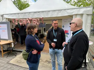 Drei Personen mit Namensschildern stehen vor einem weißen Zelt mit der Aufschrift 'Y CHATBOT' aus Luftballons