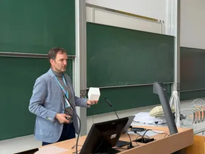 Mann mit grauem Sakko hält einen weißen Würfel vor einer grünen Tafel in einem Hörsaal