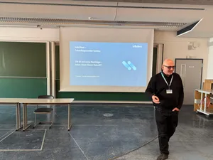 Mann mit schwarzem Pullover und Namensschild steht vor einer Leinwand mit Präsentationsfolie in einem Konferenzraum