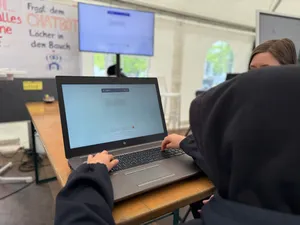 Person mit Kapuzenpullover arbeitet an einem Laptop auf einem Tisch in einem Raum mit Monitor und Plakat im Hintergrund