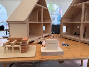 Modell eines Hauses mit offenem Dach und sichtbaren Innenräumen sowie einem kleinen Modell mit der Aufschrift 'Wärmepumpe' auf einem Tisch