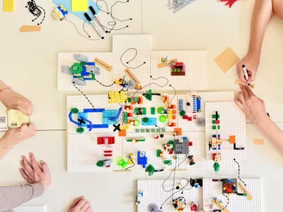 Mehrere Hände arbeiten an einer komplexen Lego-Struktur auf weißen Grundplatten mit angelegten Kabeln und beschrifteten Holzstäbchen