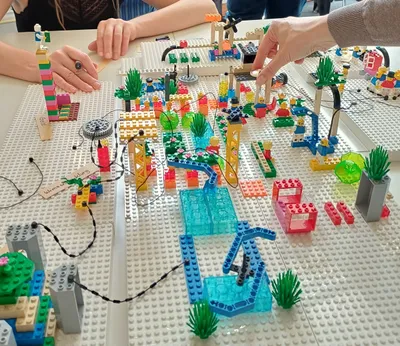 Mehrere Hände bauen auf einer großen grauen LEGO-Basisplatte eine bunte LEGO-Landschaft mit Wasserflächen, Pflanzen und verschiedenen Strukturen