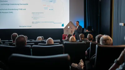 Vortragender steht vor Leinwand mit Karte und Text, Publikum sitzt in Reihen in einem Hörsaal