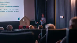 Person steht vor Leinwand mit Text und Modellhaus im dunklen Hörsaal, Publikum sitzt in Reihen
