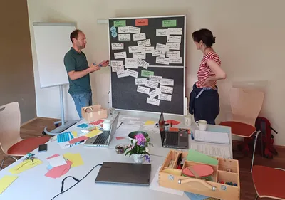 Zwei Personen diskutieren viele Karten, die an einer Pinnwand hängen.