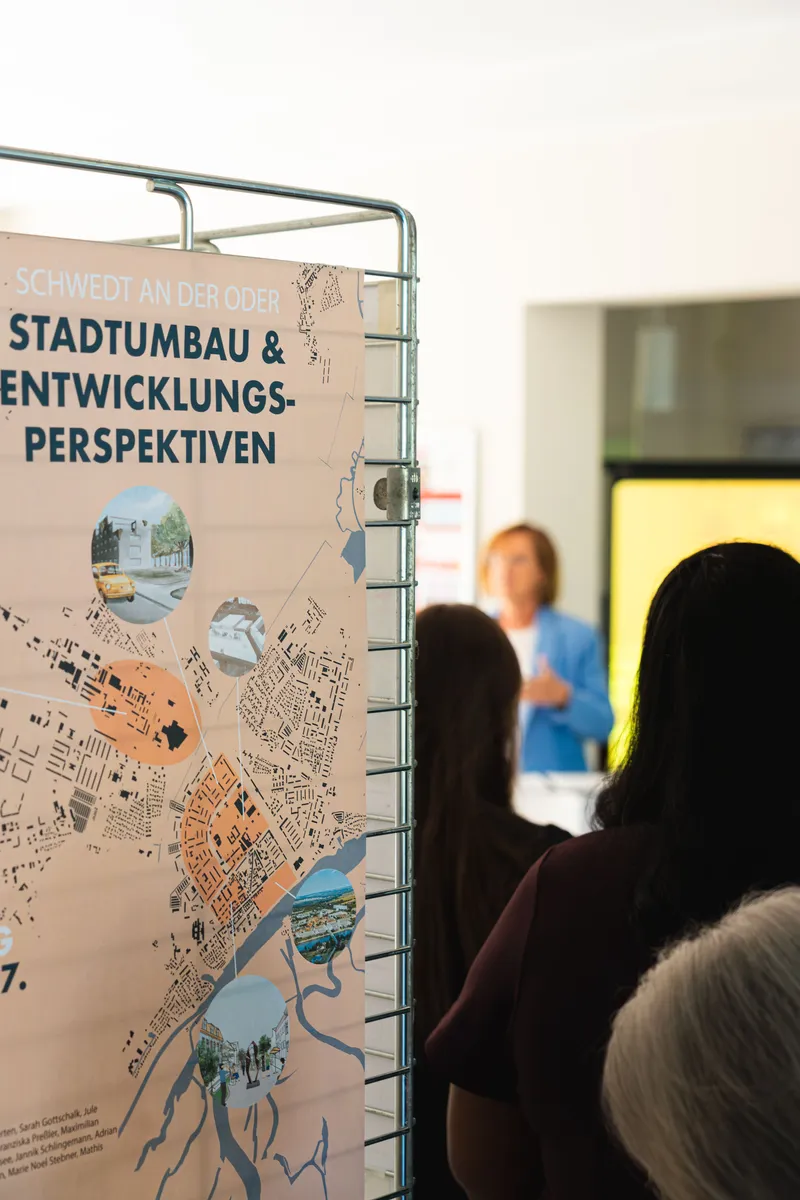 Zu Sehen ist ein Poster, im Hintergrund eine Person, die einen Vortrag hält und stehendes Publikum